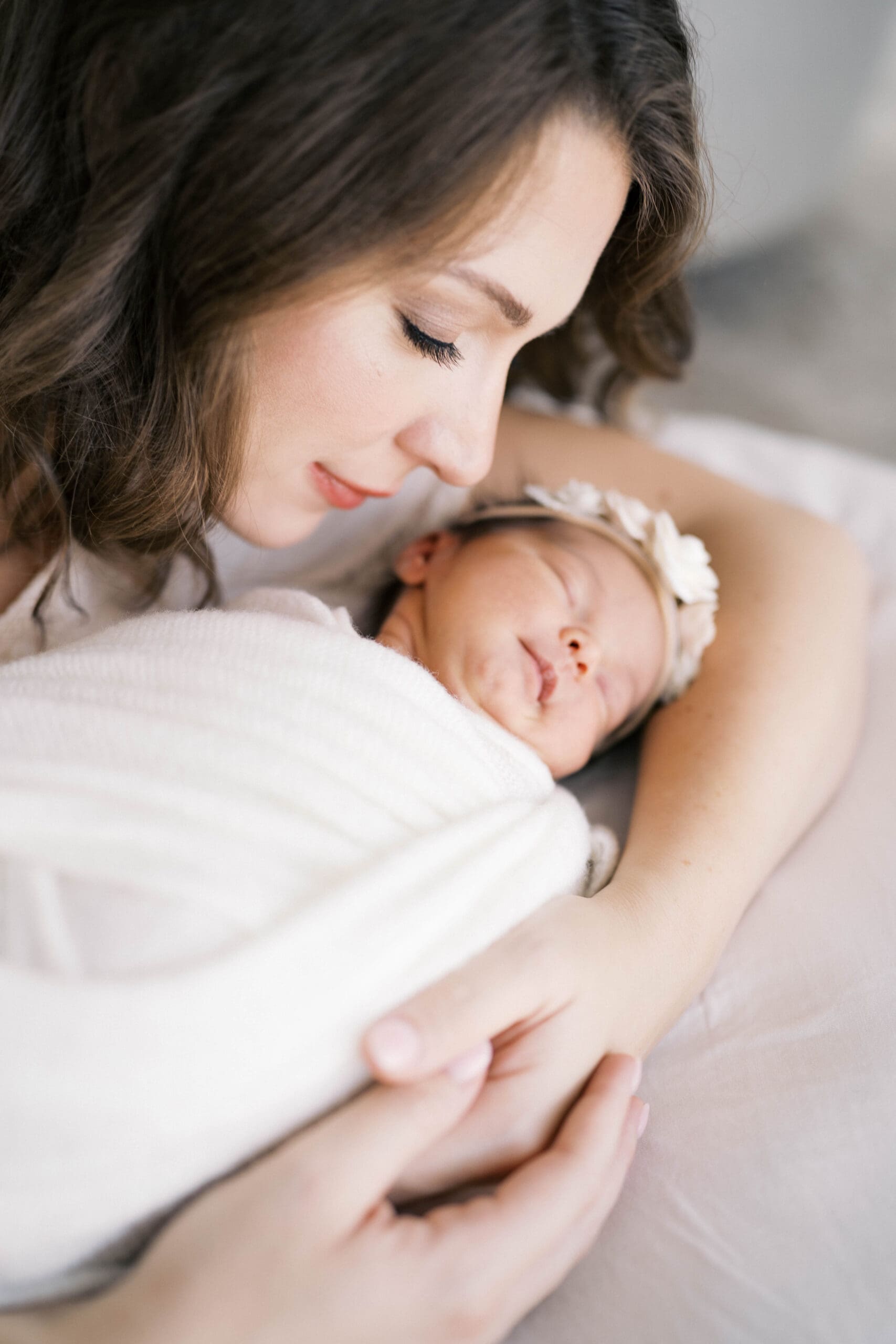 A newborn portrait of a mom lying near her baby girl in a floral bow and white swaddle shot in a natural light studio in Jacksonville, FL
