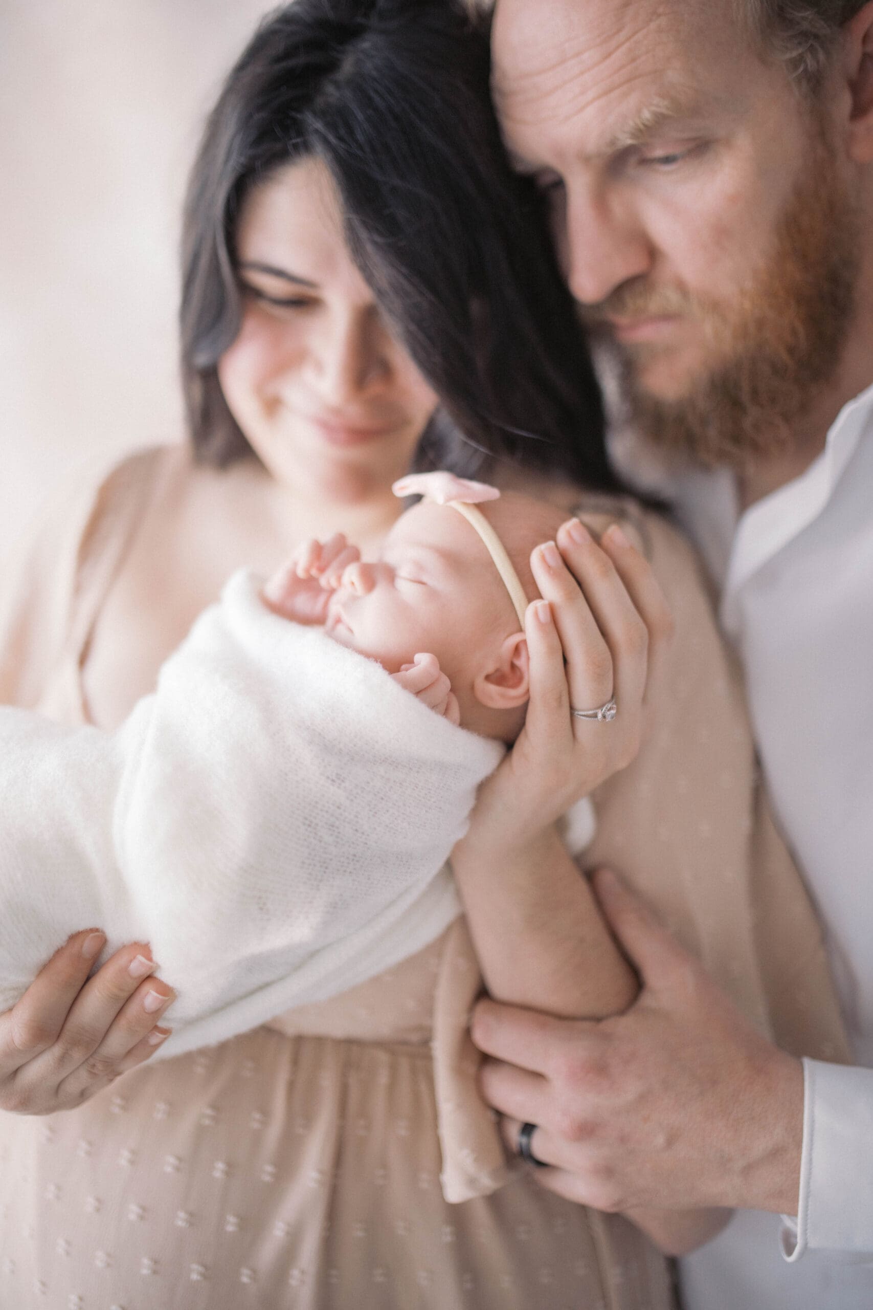 Newborn Portrait taken in a studio in Jacksonville with the mother in a pink dress holding her baby girl in a white swaddle while dad stands behind and they both gaze at their girl.