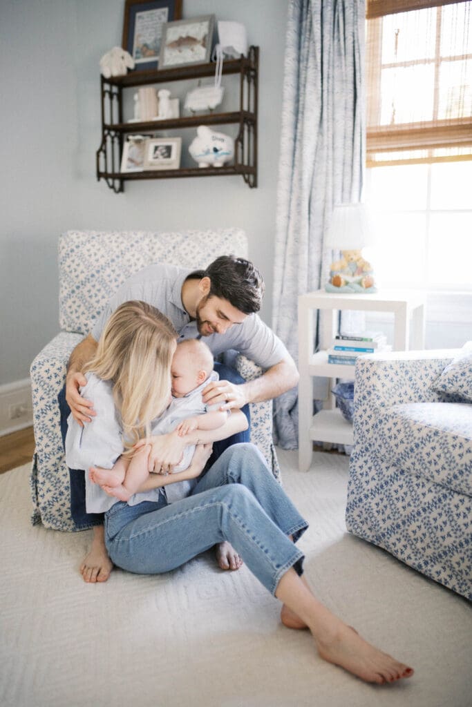Lifestyle Newborn Photo of a mom in blue jeans and dad snuggling mom while holding her baby boy in a blue bubble romper shot in a Jacksonville Home in the nursery chair