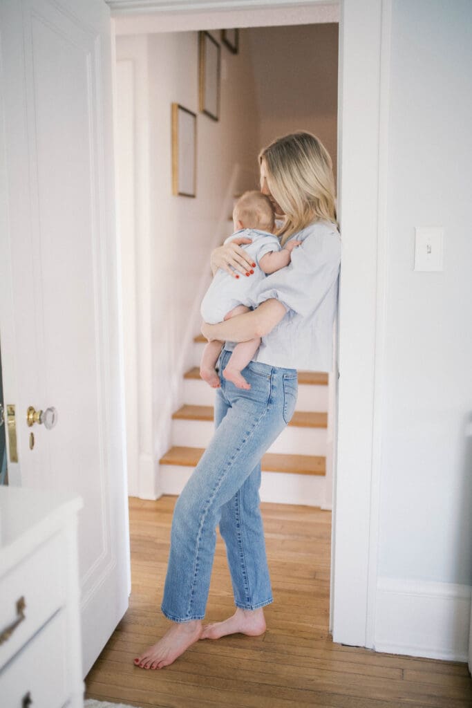 Lifestyle Newborn Photo of a mom in blue jeans holding her baby boy in a blue bubble romper shot in a Jacksonville Home by the stairs