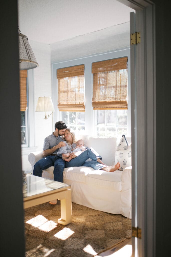 Lifestyle Newborn Photo of a mom in blue jeans and dad snuggling mom while holding her baby boy in a blue bubble romper shot in a Jacksonville Home in the sitting room on a white couch