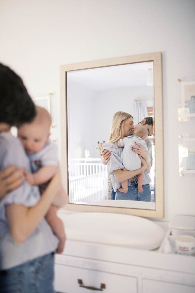 Lifestyle Newborn Photo of a mom in blue jeans and dad snuggling mom while holding her baby boy in a blue bubble romper shot in a Jacksonville Home in the nursery mirrow