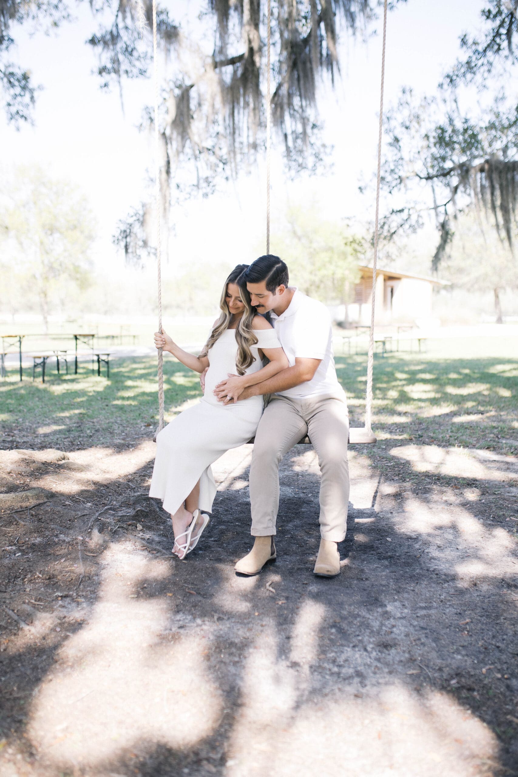 Maternity portrait of a pregnant other in a long white off the shoulder dress sitting on a large tree swing with her husband as they look down at the baby bump with a green Jacksonville park behind them