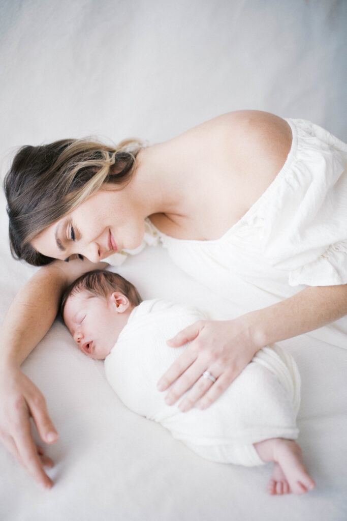 Newborn portrait of mother lying next to baby boy asleep with a neutral backdrop