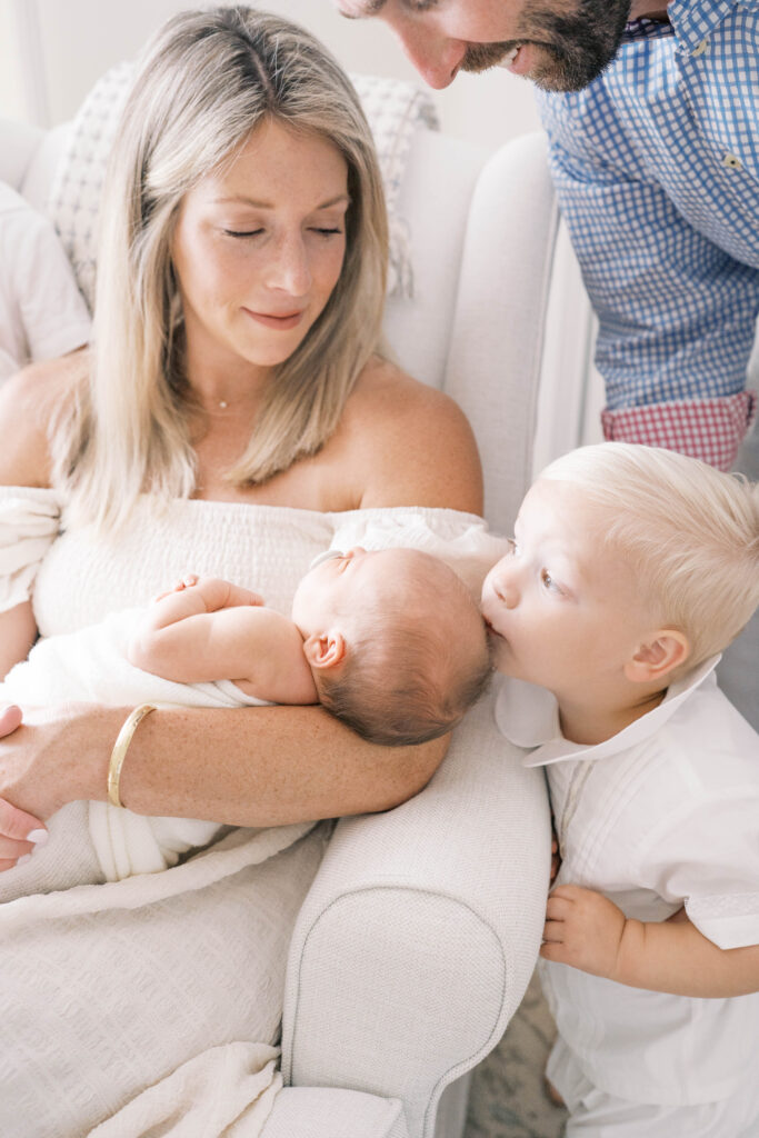 Newborn portrait of mom looking down at her middle child kissing his baby sister on her head in Jacksonville home