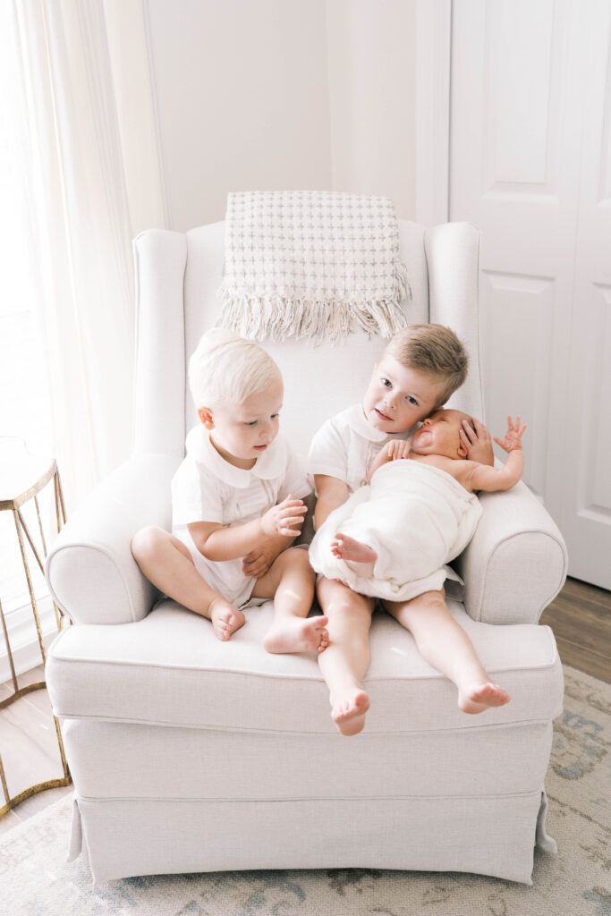 Big brothers sitting in a rocking chair snuggling their baby sister in white swaddle