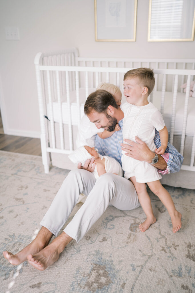 Portrait of dad sitting on the floor looking down at his baby girl as big brothers stand beside him looking at her