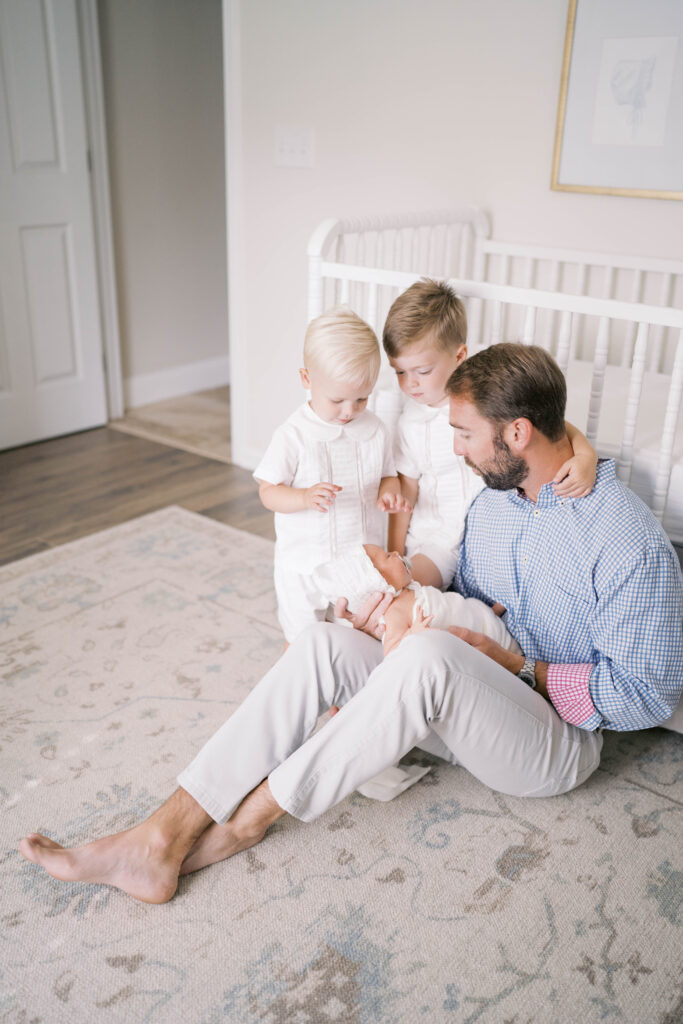 Newborn photography portrait shot in a Jacksonville home of dad holding his baby girl with his other two boys as they sit on the floor together