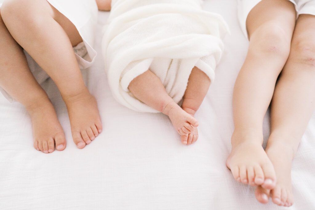 Newborn portrait of baby girl's feet peeking out of her swaddle next to her big brother's feet as they lay on mom and dad's bed during a photography session in Jacksonville