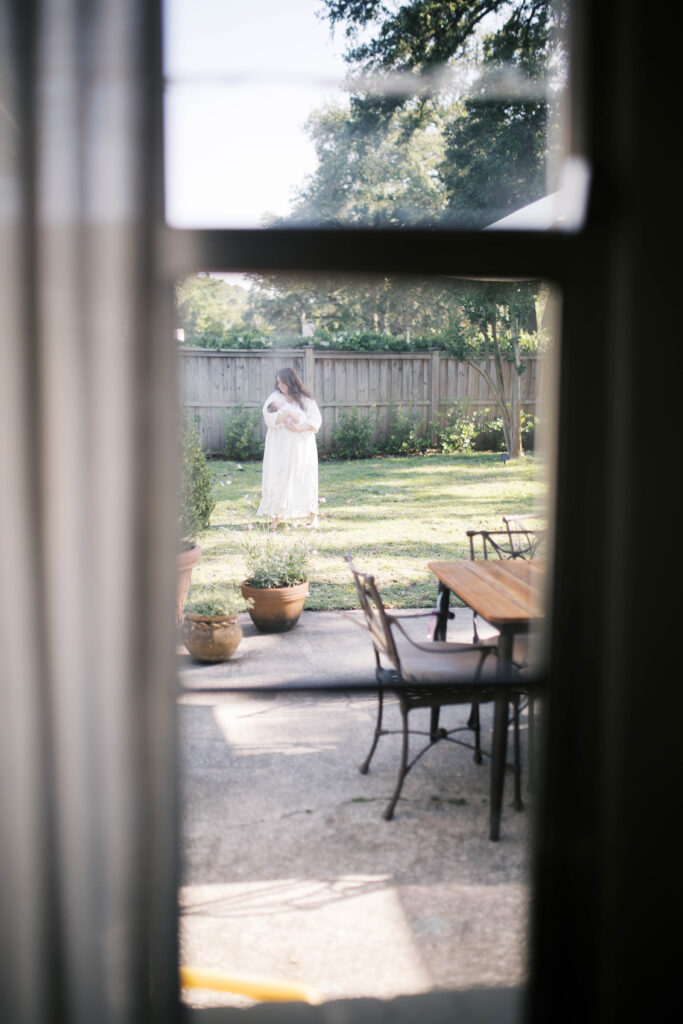 newborn portrait shot through an antique window of a mom swaying her baby girl in her backyard