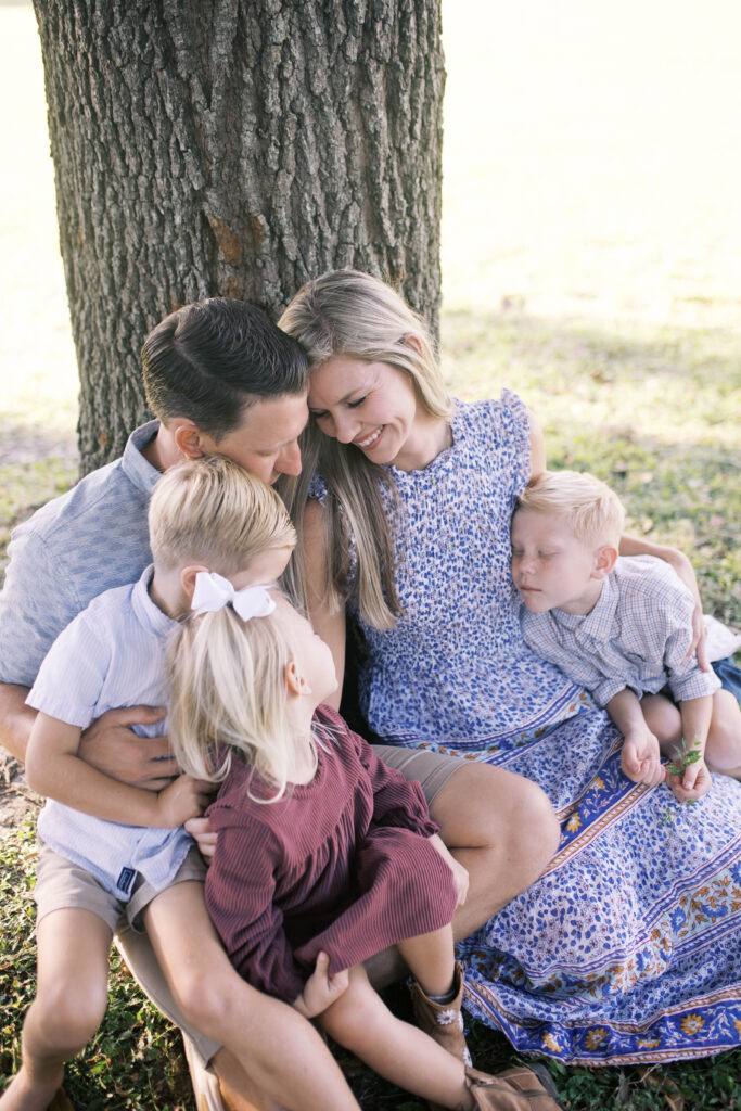 mom and dad seated against a large oak tree at a park in Jacksonville FL with two boys and toddler girl sitting across their laps snuggling and smiling together