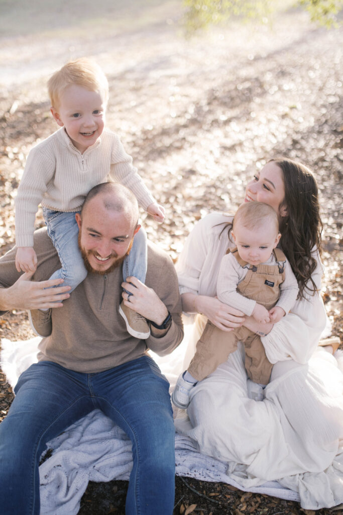 A mom and dad seated on the ground at a park in San Marco with todler son on dad's shoulders and baby boy in mom's lap as they all laugh