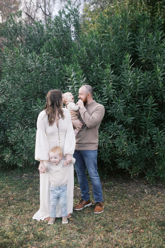 Portrait of mom holding toddler son's arms as dad plays with baby boy in front of a large green leafy tree in Jacksonville FL