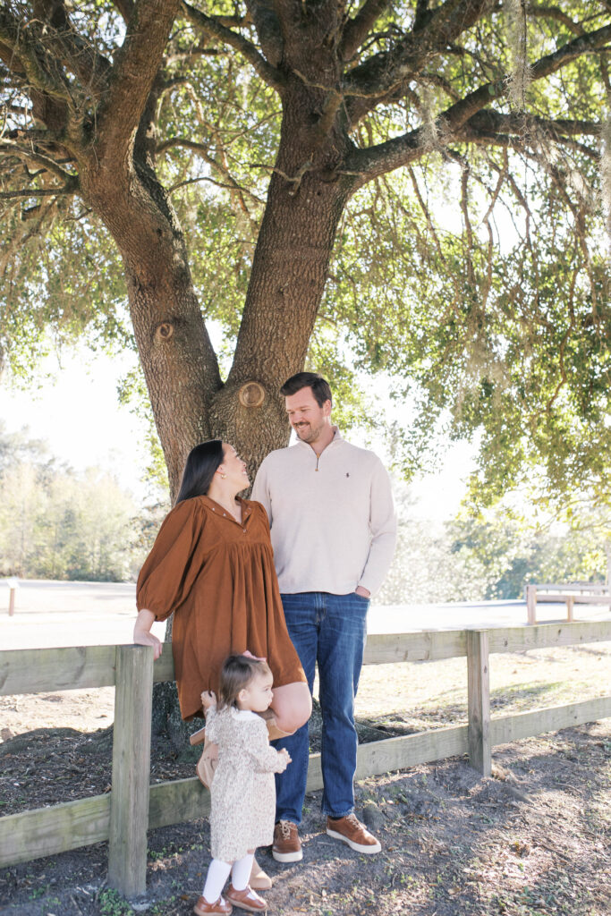 Portrait of family of 3 with parents leaning against wooden fencing in front of a large oak tree as toddler daughter looks out into the sun and parents are smiling