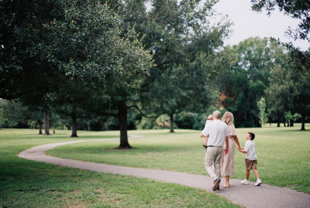 Family photo of mom dad and two sons holding hands and walking away from the camera on a winding sidewalk surrounded by green trees in a park in San Marco